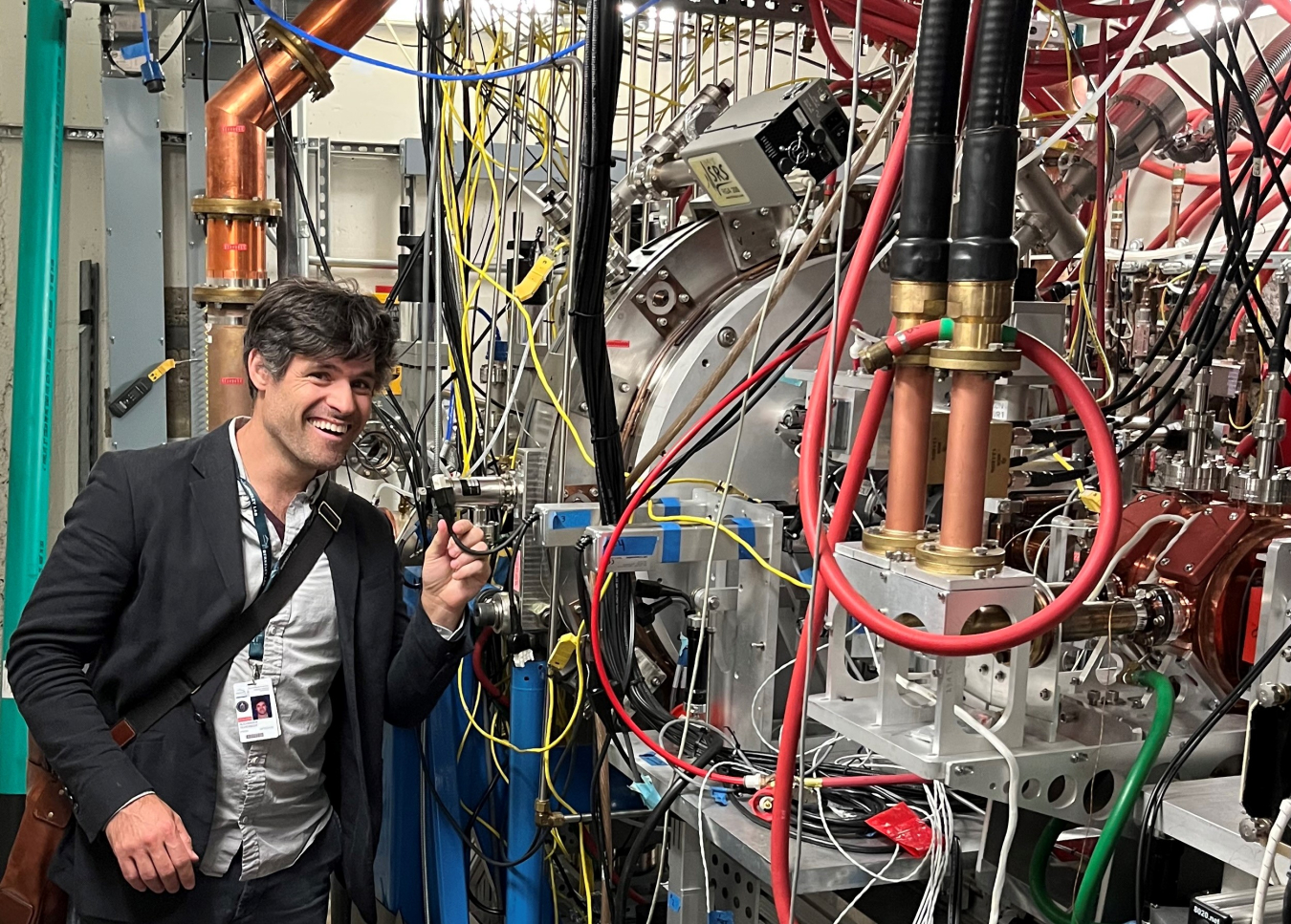 Alexander Scheinker stands next to a section of particle accelerator machinery that has loops of wires of various colors hanging from it. 