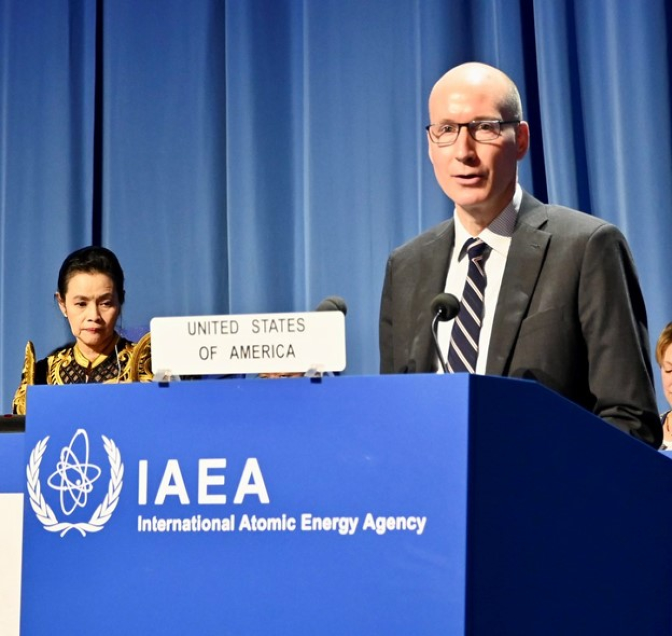 A man gives a speech at a podium that says IAEA: International Atomic Energy Agency. A sign on the podium says United States of America.