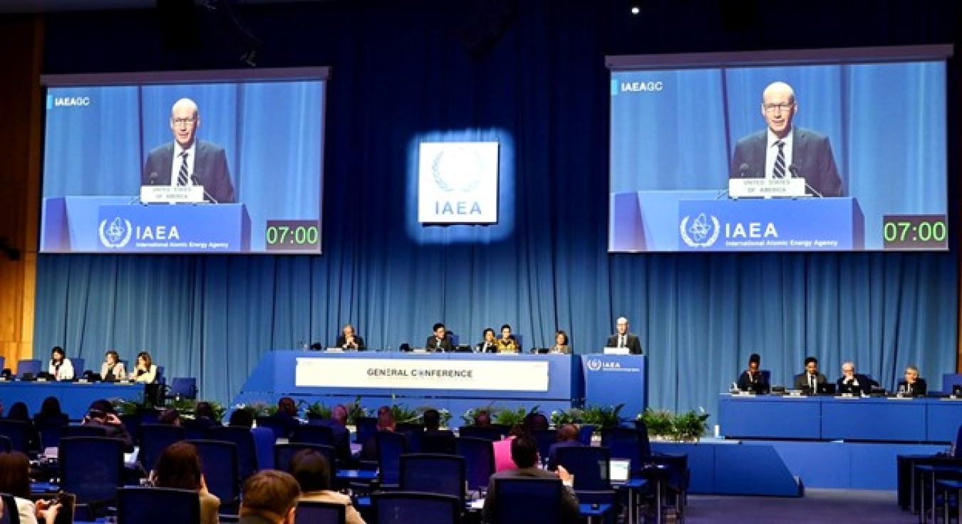 A man presents from a podium to a large audience. There are two large screens behind him showing the podium, as well. The words International Atomic Energy Agency are at the front of the room.