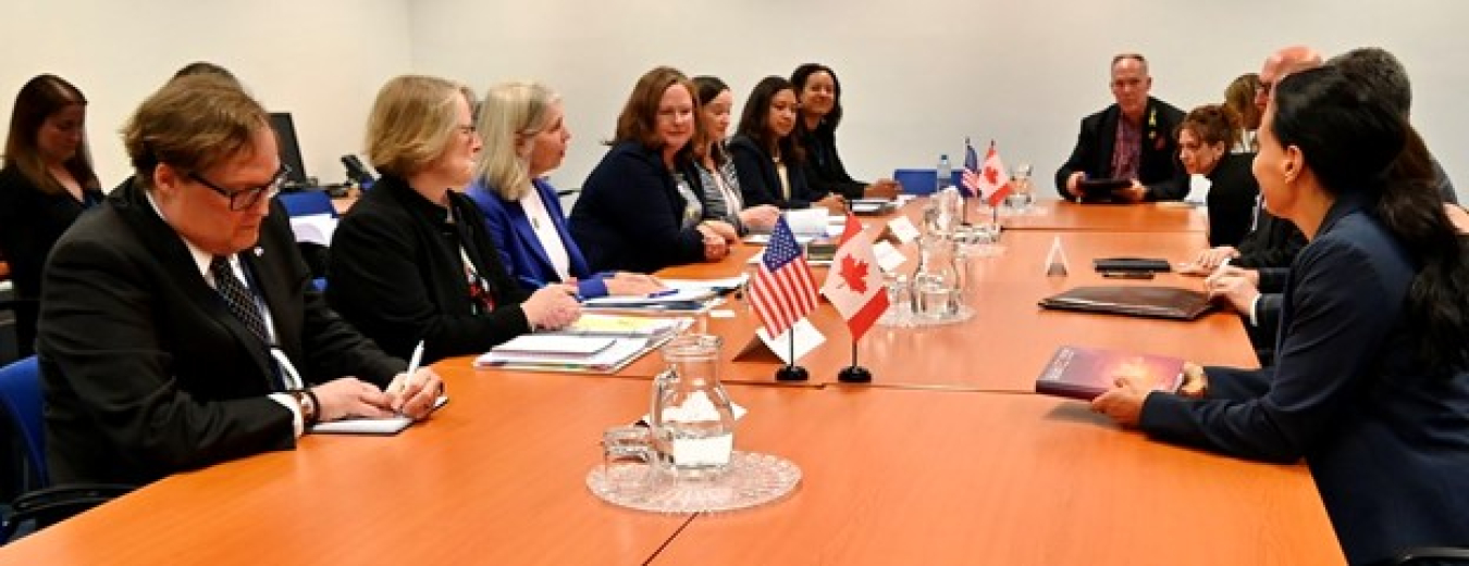 Two groups of people face each other on the sides of a large conference table. Small U.S. and Canadian flags sit in a stand between them.