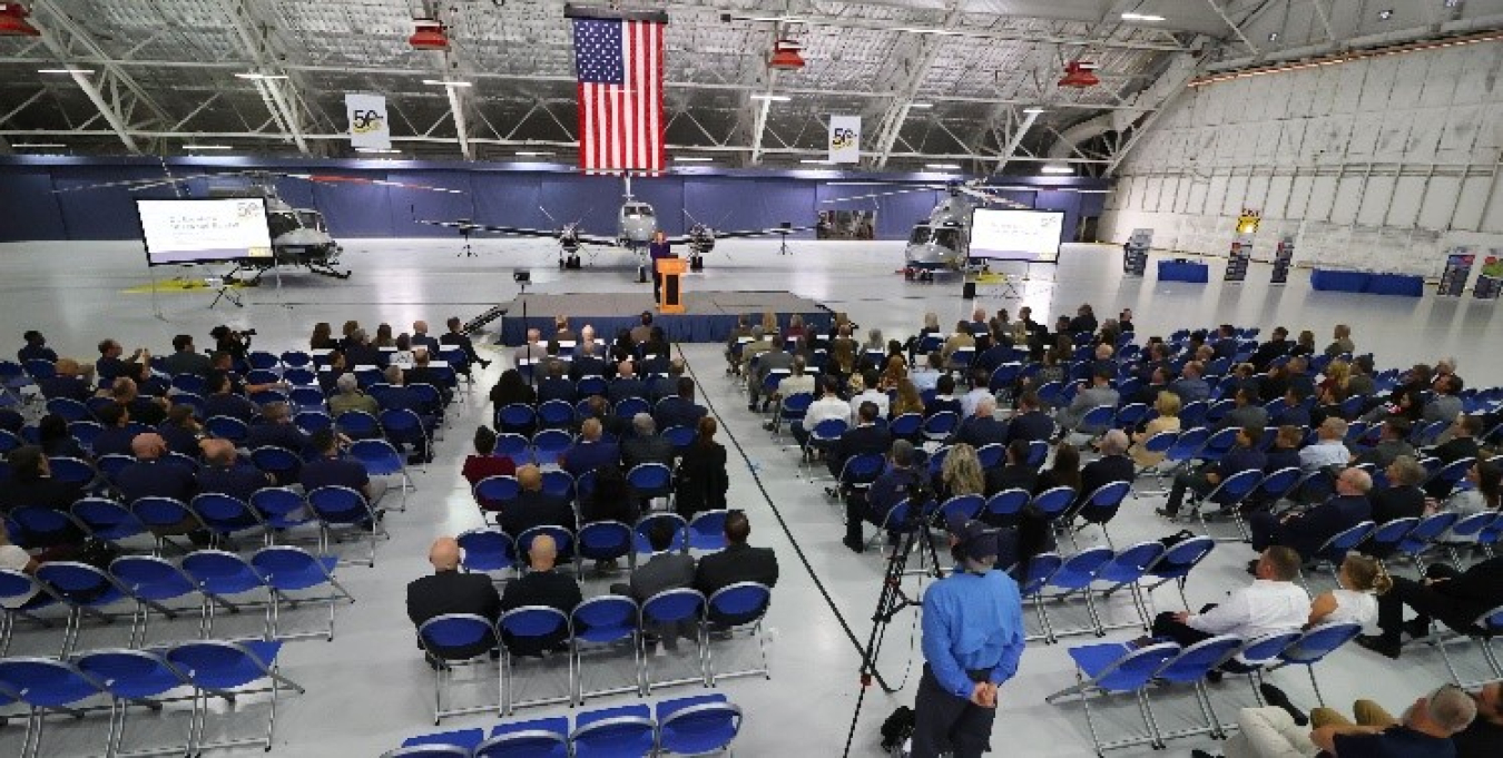 A hundreds of people sit in rows in a large hangar. American flags hang from the ceiling. Aircraft are parked behind a podium where a person is speaking.