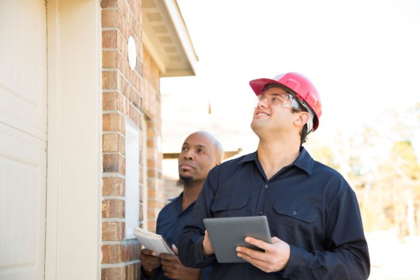 Two men, one wearing a hard hat, inspecting the exterior of a brick home.