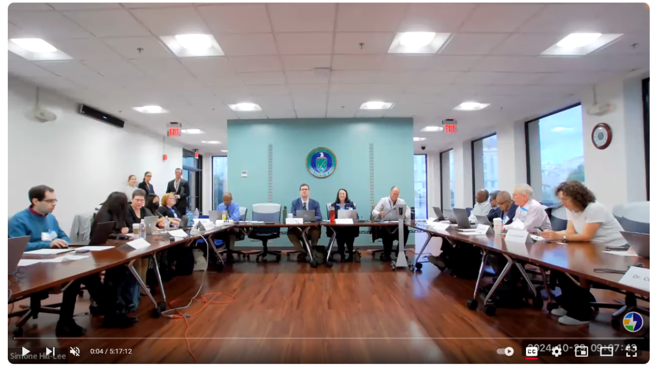 A screenshot of a video showing people sitting at a table in front of a government seal.