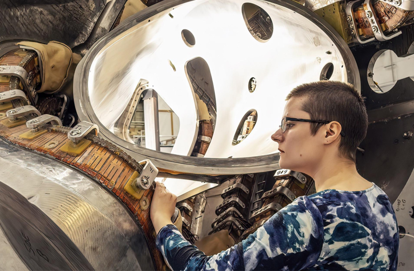 Alexandra LeViness (a white woman with a blue and white patterned long-sleeved shirt on), with her hand on a metal device much larger than her. A metal piece made of thick silver-colored metal with a ribbed copper-colored belt-like section snakes up and away from her, above her head.