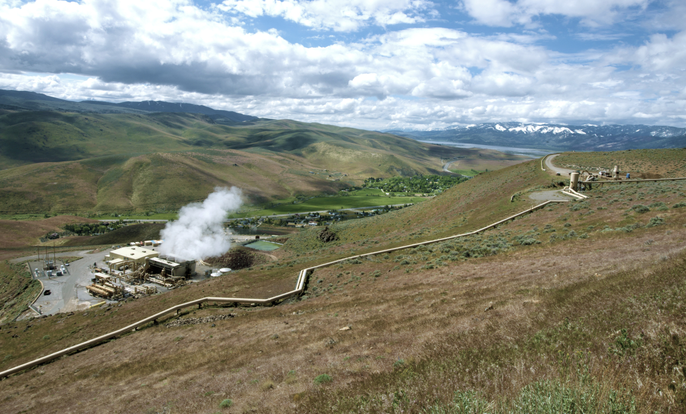 A geothermal plant, which looks like a series of pipes and tubes with steam rising from them, sit in hills with rolling hills extending back into mountains on the horizon.