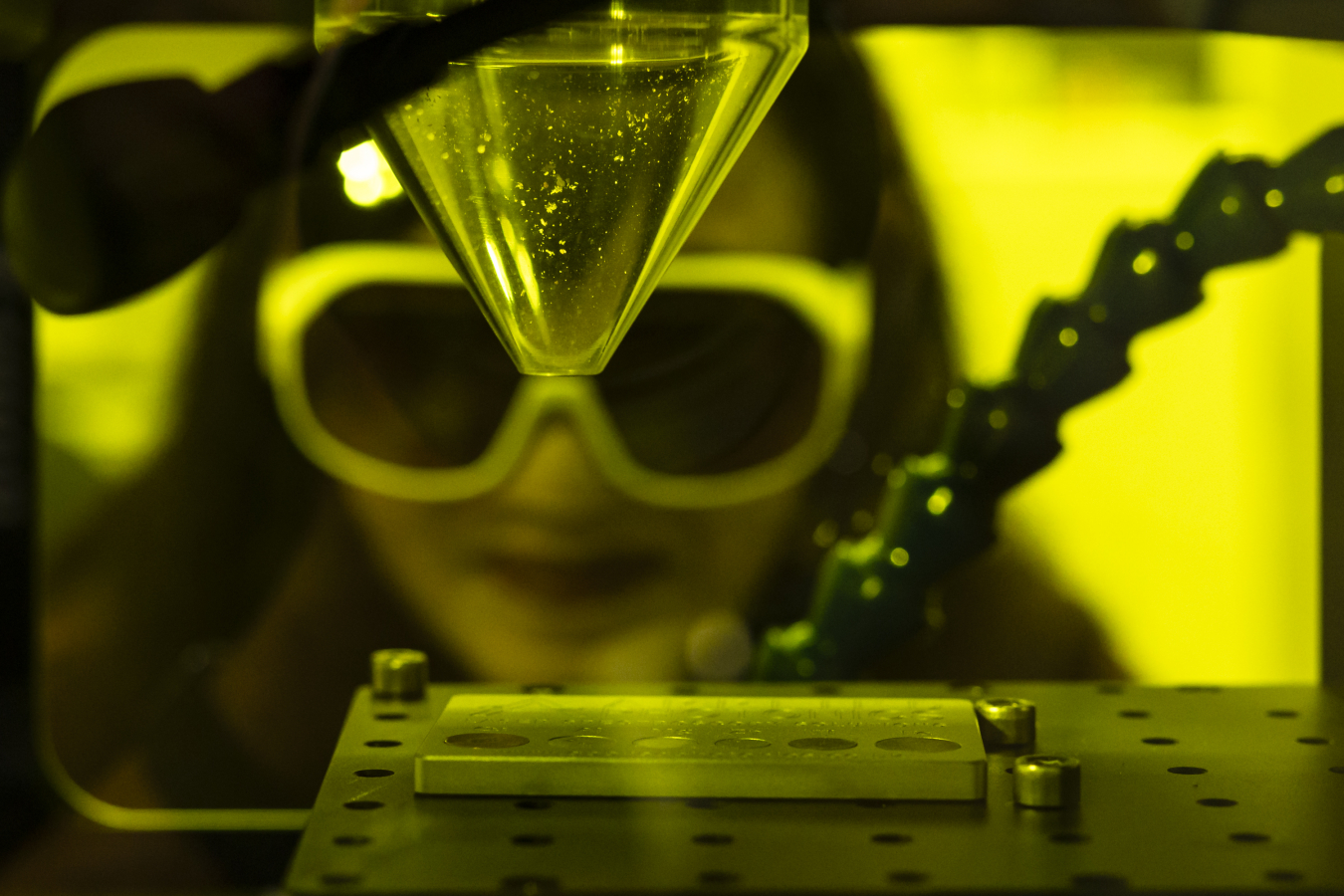 Madhavi Martin (a Southeast Asian woman with large lab goggles on) looks into a yellow-tinted box used for laser-induced breakdown spectroscopy. Inside the box is a clear funnel that is pointed towards a metal sheet with holes in it.