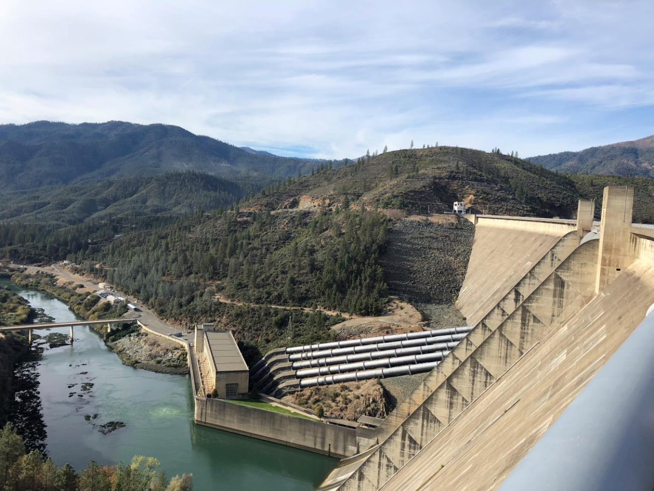 Side view of a large hydropower dam with the outflowing river and tree lined valley in the background