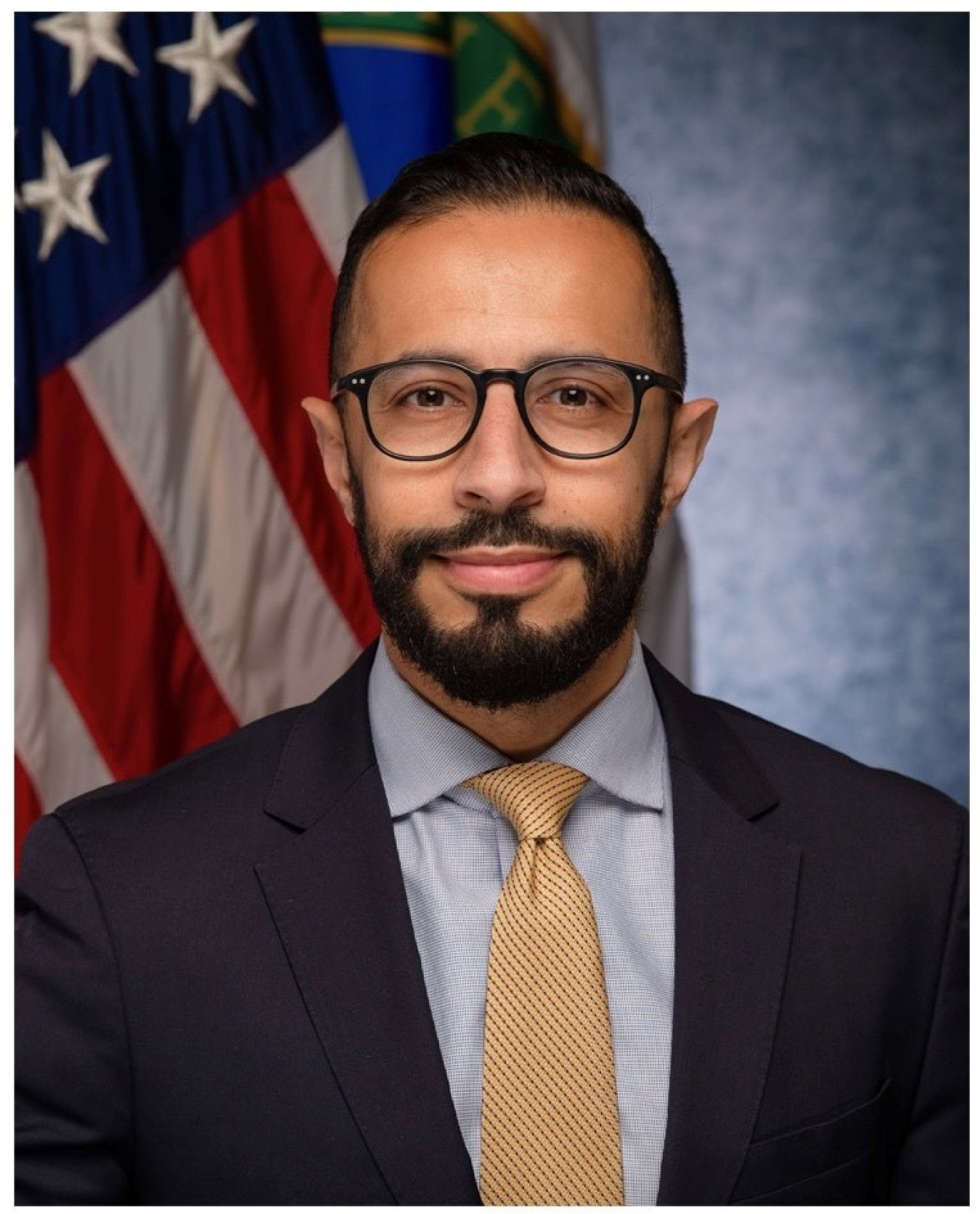 Headshot of Dr. Rian Bahran, showing him wearing a suit and tie with an American flag behind him.