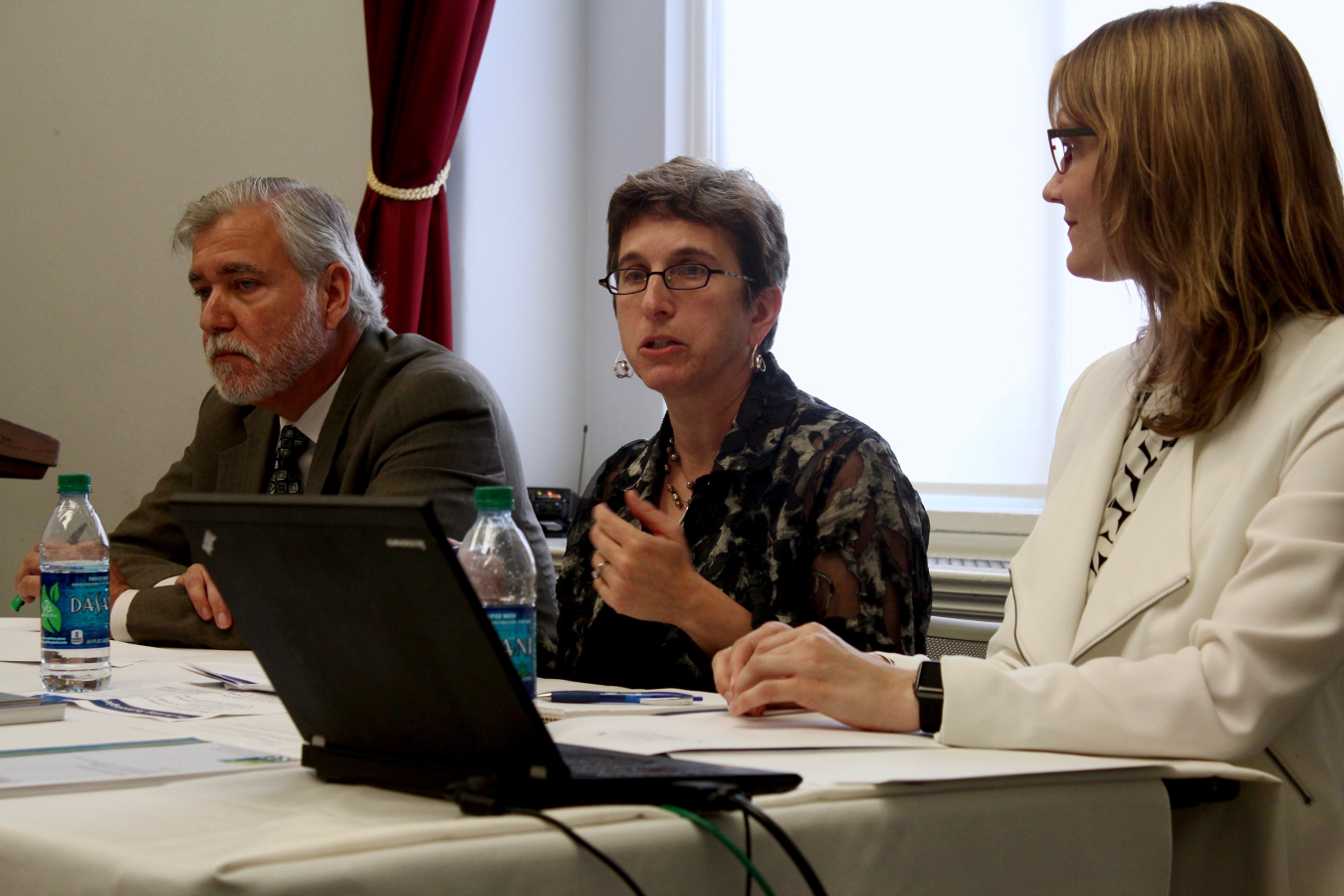 Three people sitting at a table in a meeting, with the focus on the woman in the center.