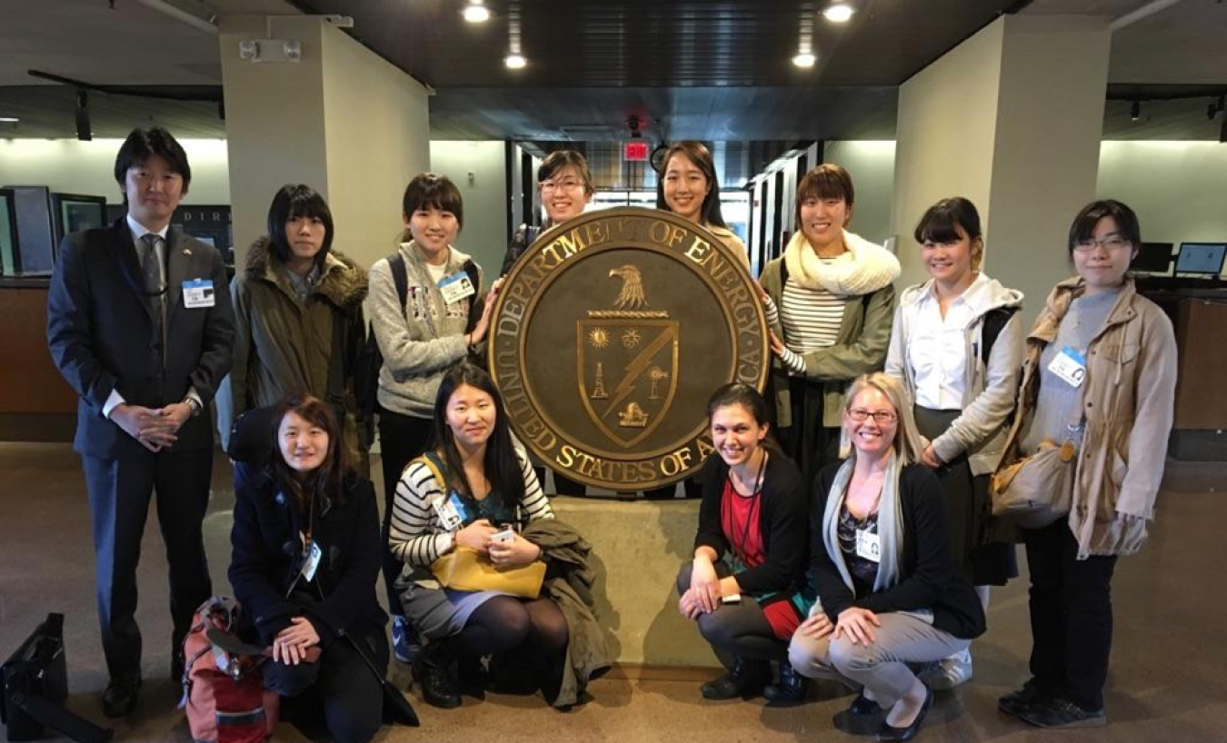 Photo of a group of people facing the camera as they stand around the sculpture of the Department of Energy seal in the DOE headquarters lobby.