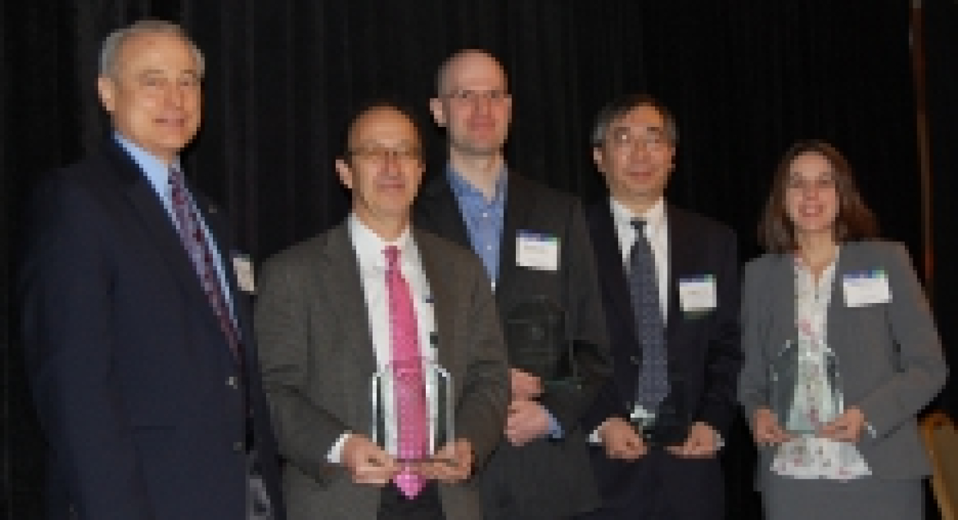 Photo of five people standing and facing the camera. Four people are holding award trophies.