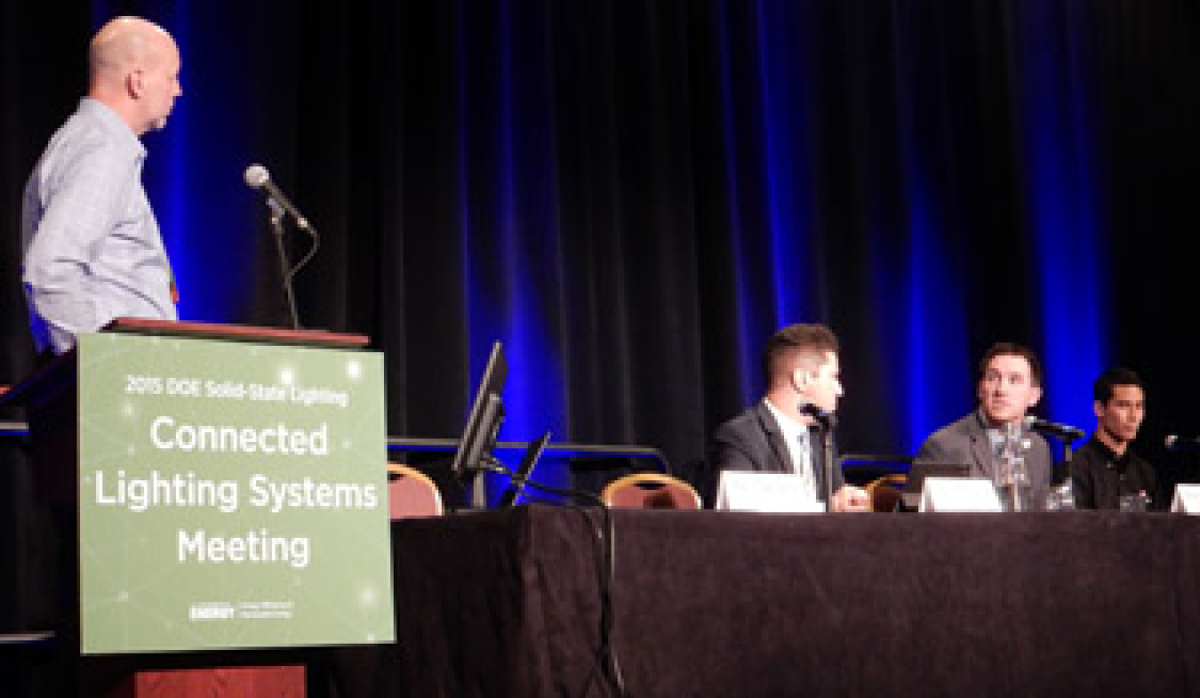 Man standing at a lectern, looking toward the table next to the lectern that has three people seated at it, all in front of an audience.