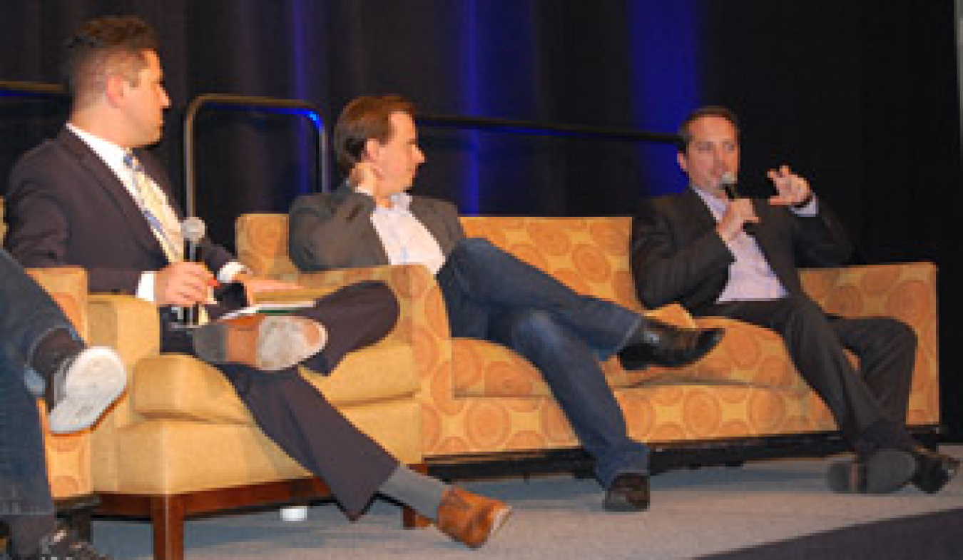 Three men in business suits seated on couches, addressing an audience during a panel.