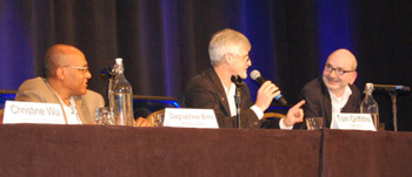 Three men seated at a table with microphones addressing an audience during a panel discussion.