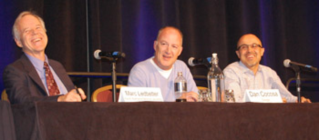 Three men seated at a table with microphones to address an audience during a panel discussion; the men are laughing.