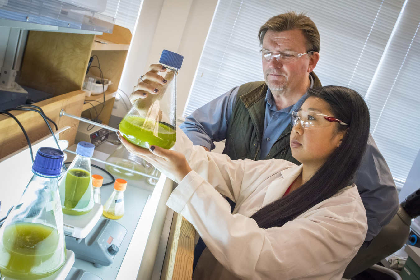 Photo of a female student scientist holding up a bottle of algae while a teacher looks on.