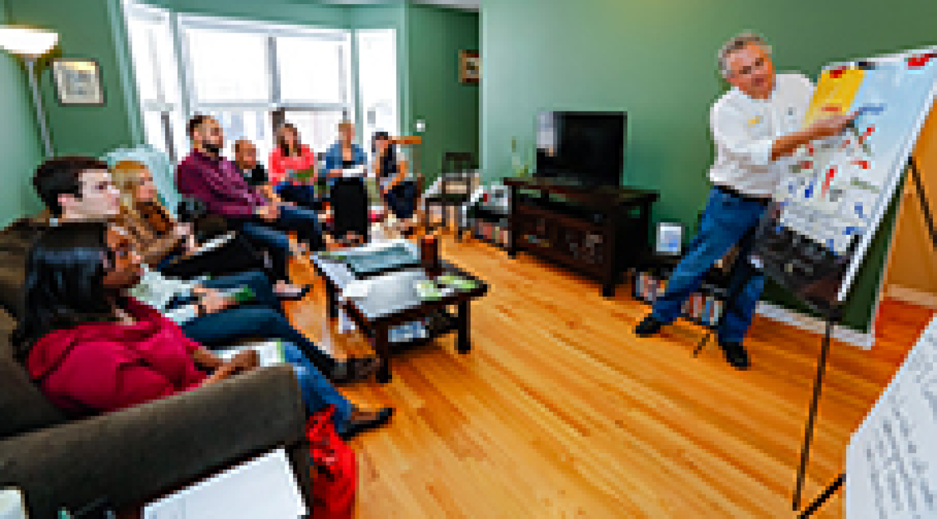 A group of people seated in a casual living room scene, focused on a man presenting to them with a flipchart.