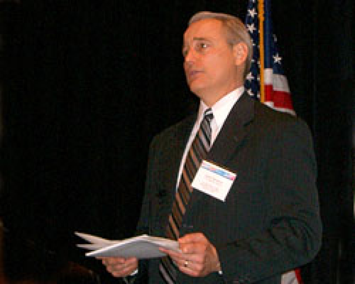 Photo of man standing, a name tag on the lapel of his suit, holding sheets of paper, with the American flag behind him.