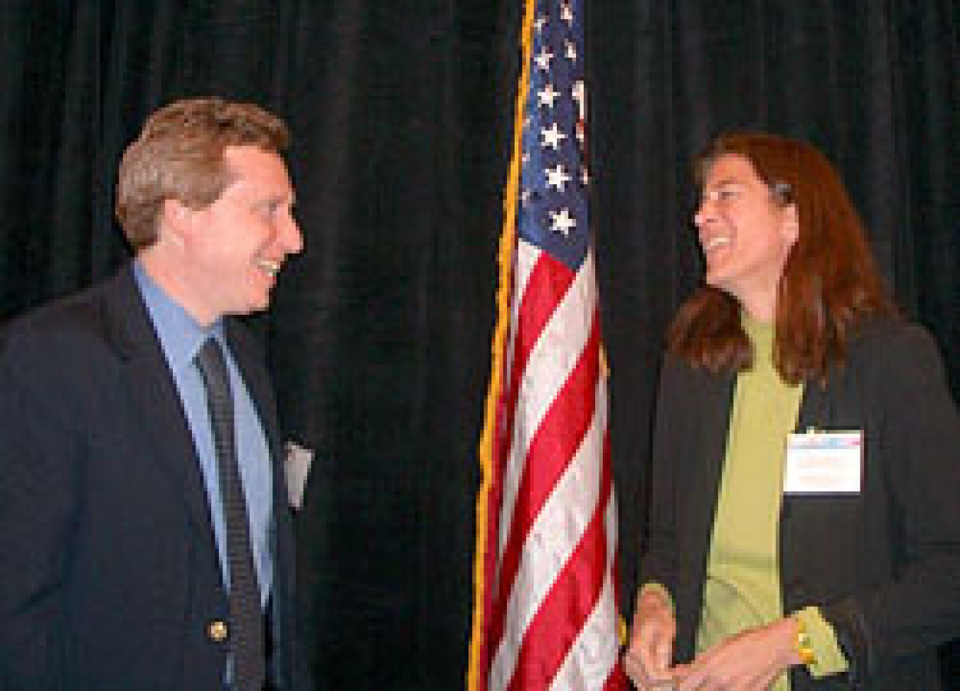 Photo of a man on the left and a woman on the right, standing, facing each other, smiling/laughing, with the American flag between them slightly in the background.