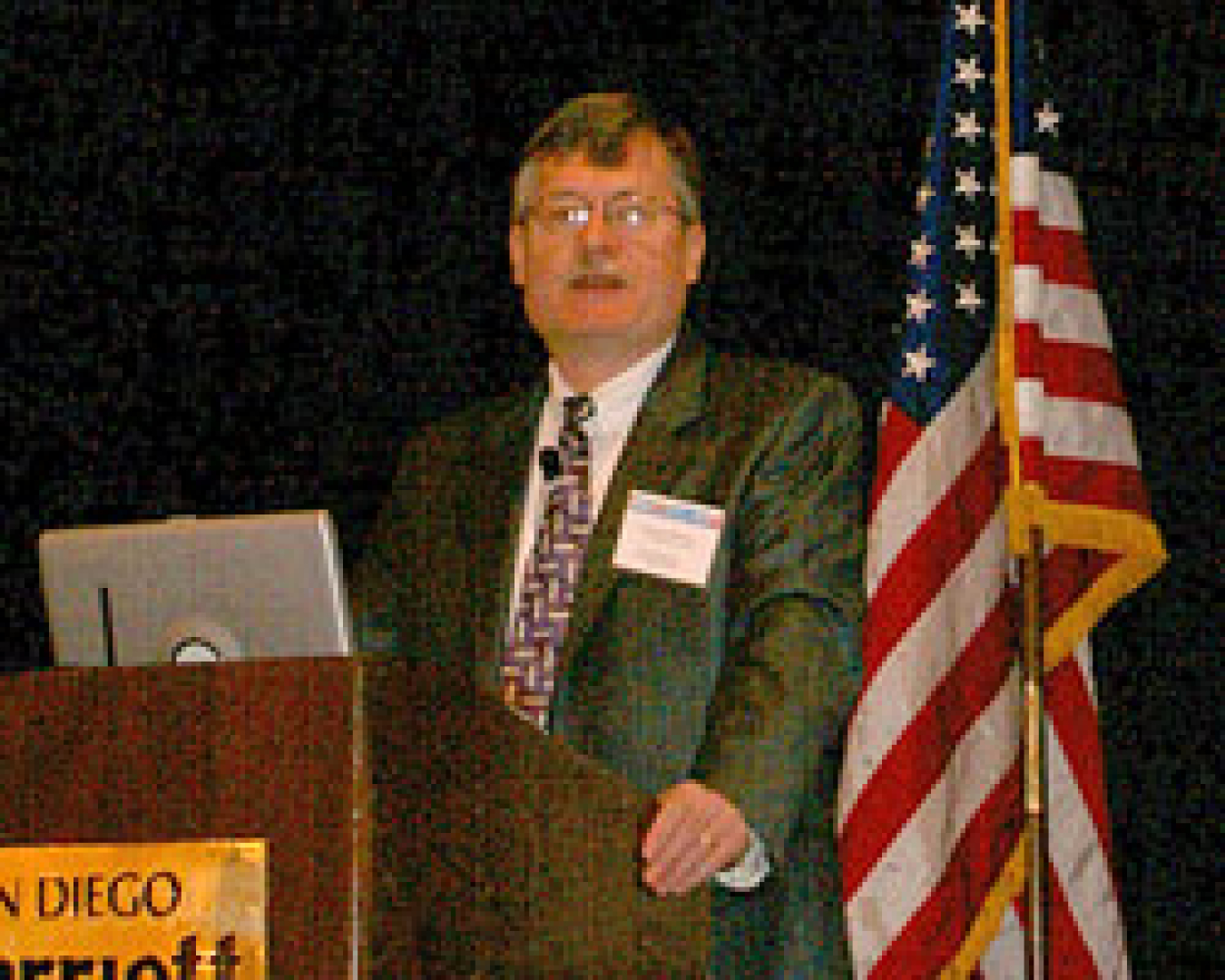 Photo of a man in a suit standing behind a podium, with the American flag on the right.