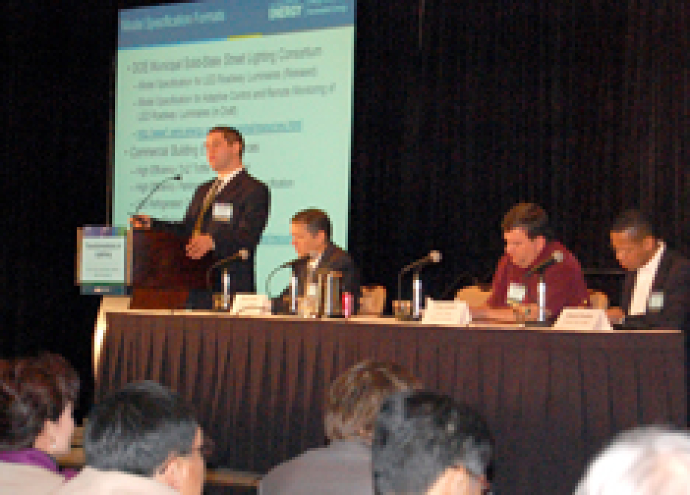 Photo of three men sitting at a table with microphones on it. Another man is standing at a podium in front of a projection screen.