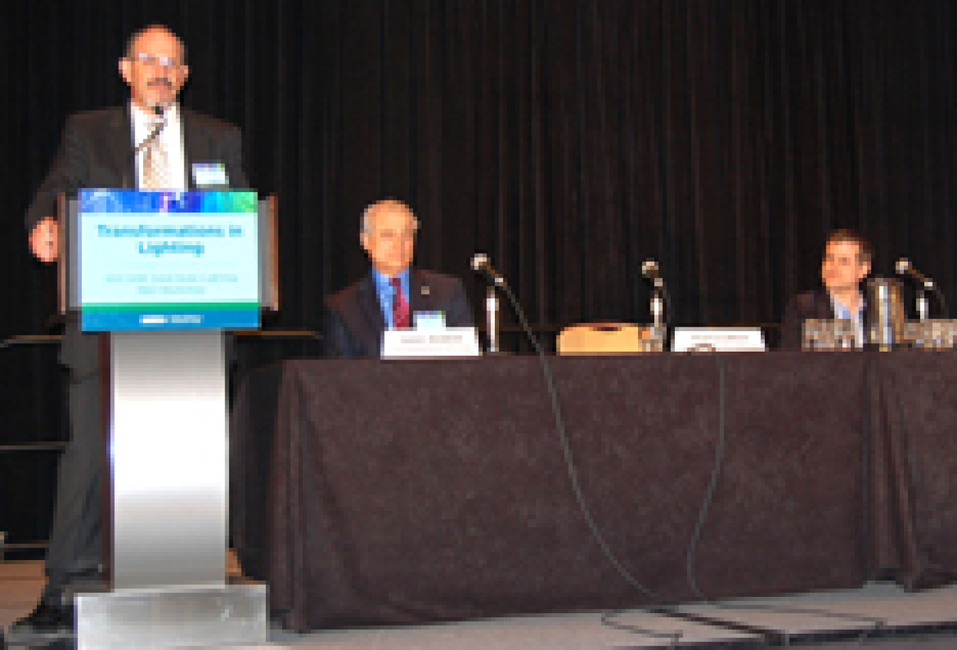 Photo of two men sitting at a table with microphones on it. A third man is standing at a podium.