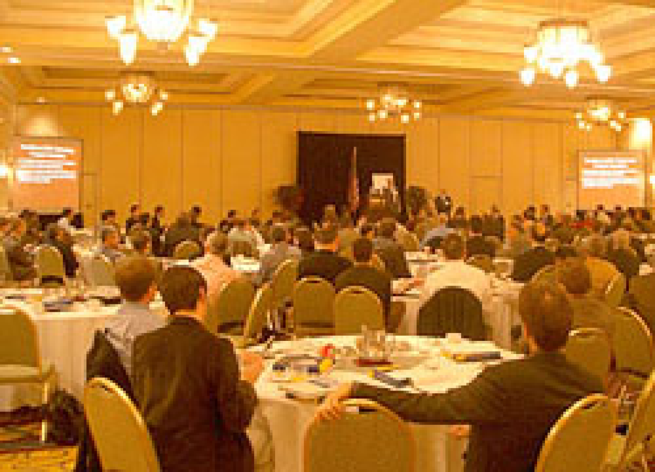 Photo taken from the back of a large ballroom showing the backs of a large crowd seated viewing a presentation on large screens to the right and to the left, and a speaker in the distance in the center standing at a podium next to the U.S. flag.