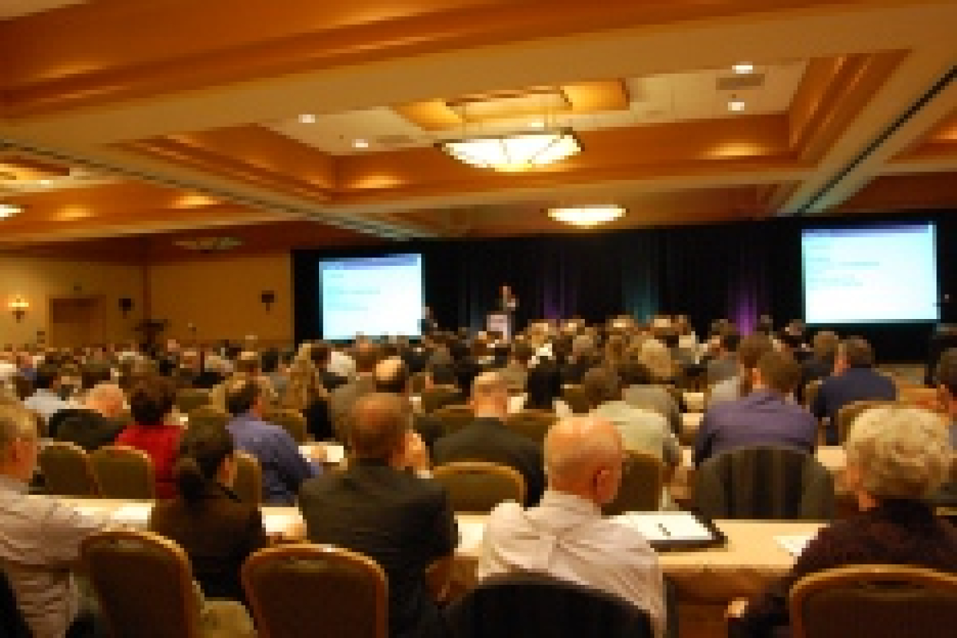 Photo of a large group of people sitting at tables, facing a screen and speakers at the front of the room.