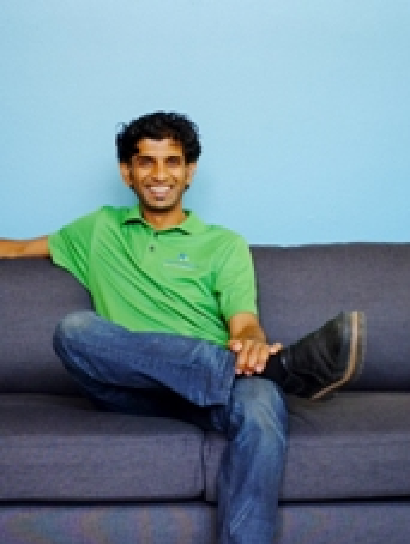 Photo of a young man sitting on a couch, looking at the camera, dressed in jeans and polo shirt.