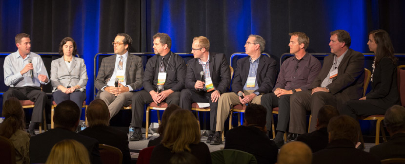 Group of nine people in business attire sitting in a row of chairs, at the head of a room, addressing an audience.