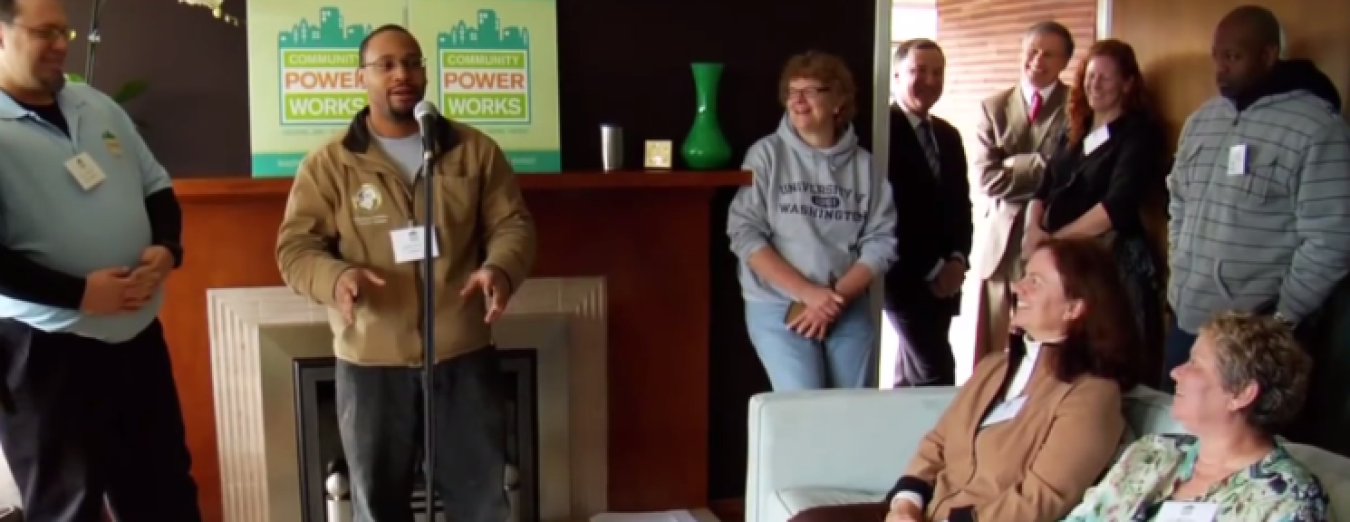 Photo of a small group of people in the living room of a home listening to a man speaking at a microphone.