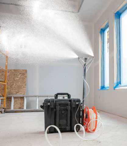 Photo of room with a black box on the floor. A pole rising up from the box has two nozzles at the top spraying a white substance into the room.